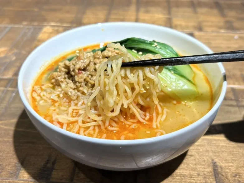 調理した担々麺を白い器に具材と一緒に盛り付け、箸で麺を持ち上げる画像