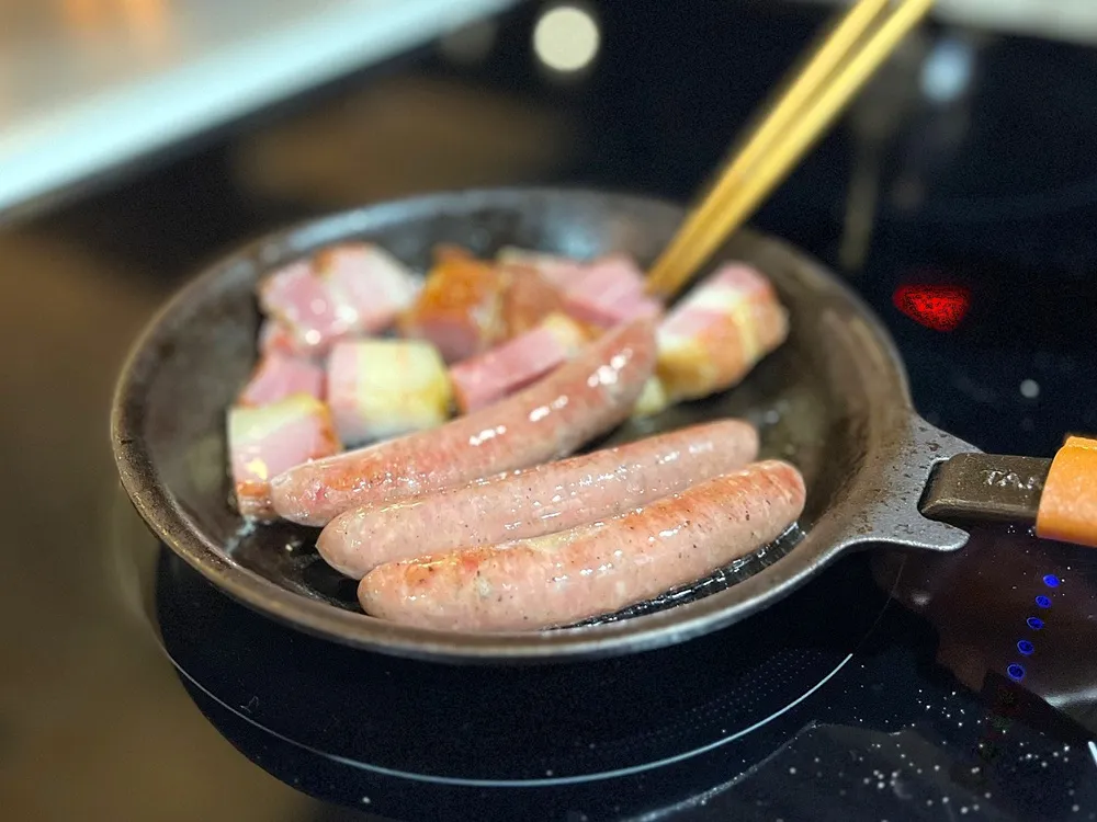 鉄製のフライパンでソーセージとベーコンを焼いている画像